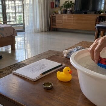 Hands lower a small plastic boat into a shallow basin of water beside a rubber duck, bottle cap, notebook and school shoes.